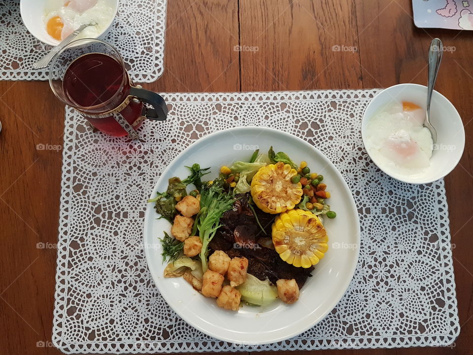 Top view of a complete full meal of meat served with vegetables on white plate with place mat and bowl of half boiled eggs set on wooden table with a drink