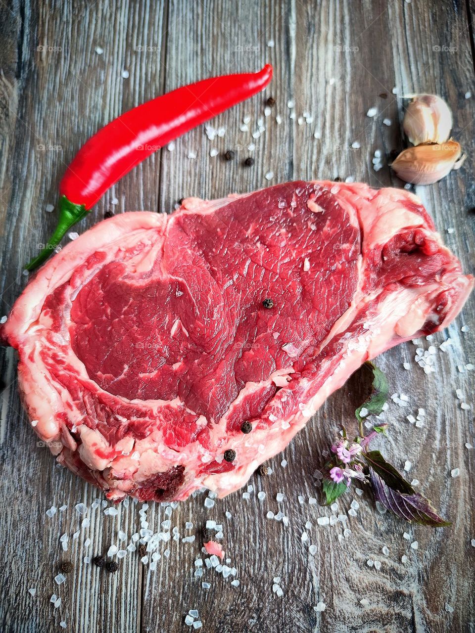 Healthy food.  Meat is a source of natural amino acids.  On a wooden surface is a piece of beef steak, red chili pepper, white salt, peppercorns, garlic cloves and a sprig of basil