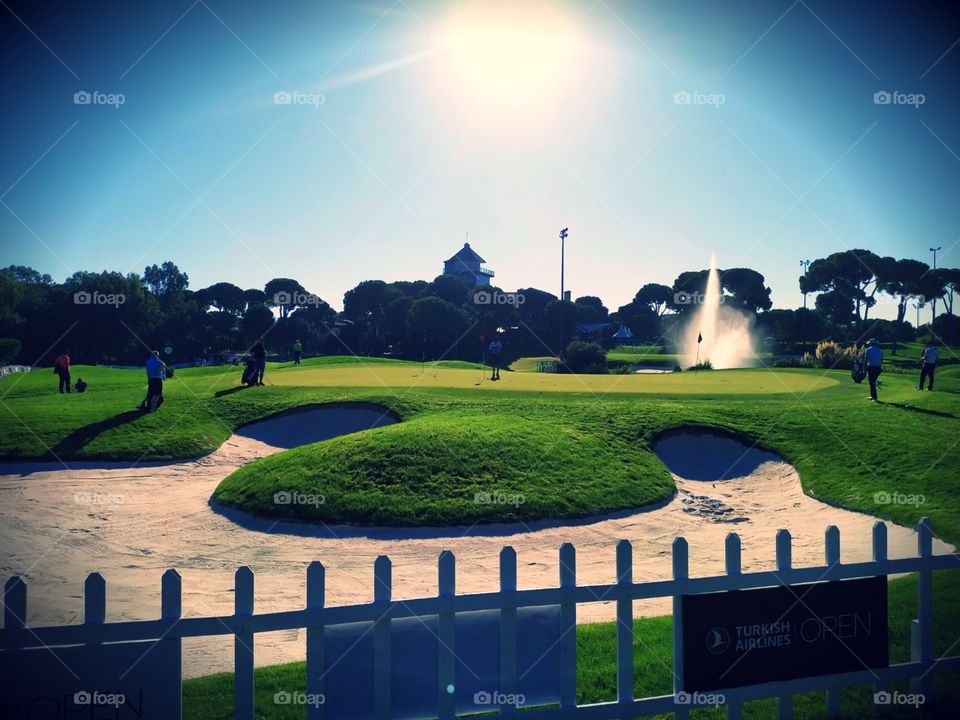 A beautiful golf course view with huge bunker, nice green, trees, water fountain and players. With beautiful blue sky and sunlight.