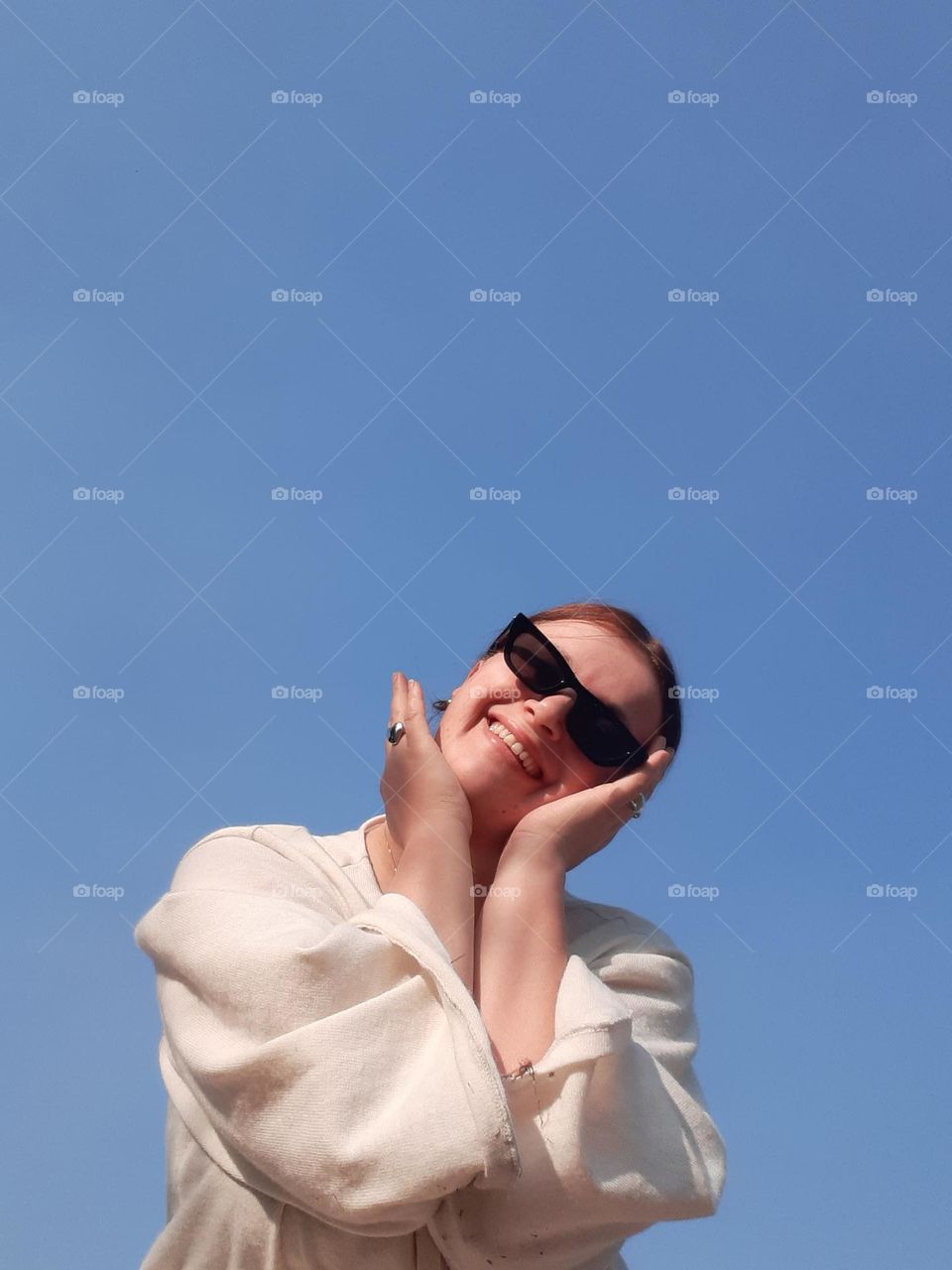 comedy female portrait of fair-skinned girl in sunglasses smiling against blue sky and holding hands near face