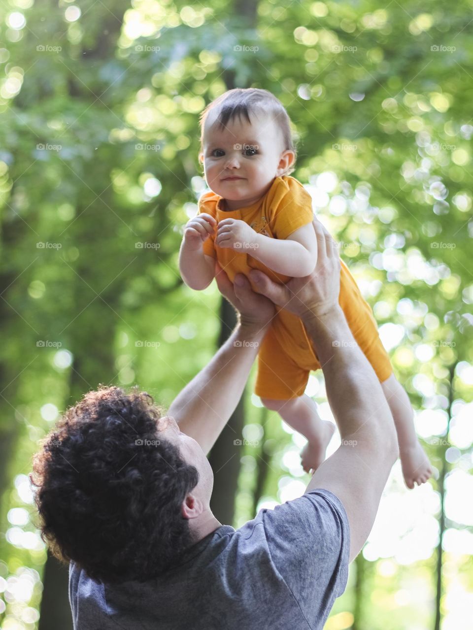 Portrait of a young caucasian man with curly brown hair in a gray t-shirt from the back throwing up his little daughter in a yellow bodysuit in the forest on a clear sunny day on a blurred tree line, vtd side close-up. The concept of dads, a walk in