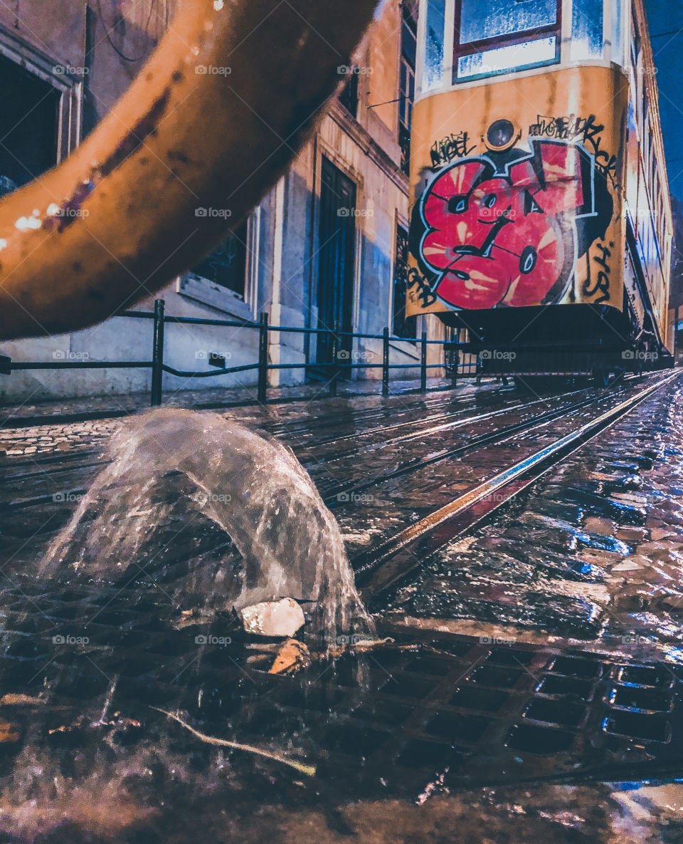 Overflowing water spews from the ground, nearby an ascending tram on a rainy night in Lisbon