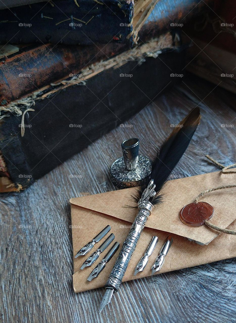 On the wooden one lies a brown envelope with an official wax seal.On the envelope is a metal fountain pen with a bird feather. Next to the envelope are metal nibs with different tips.Near the envelope is a metal inkwell.Old books in the background