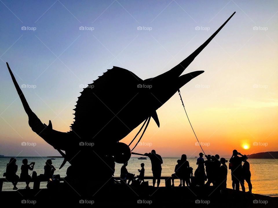 Stunning magic dramatic sunset over the smooth sea water surface with the dark silhouette of the peoples and giant symbol of Ao Nang marlin fish statue 